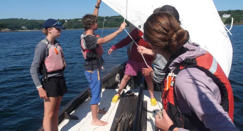 A group of students wearing life jackets adjust a white sail on a boat on blue water. 